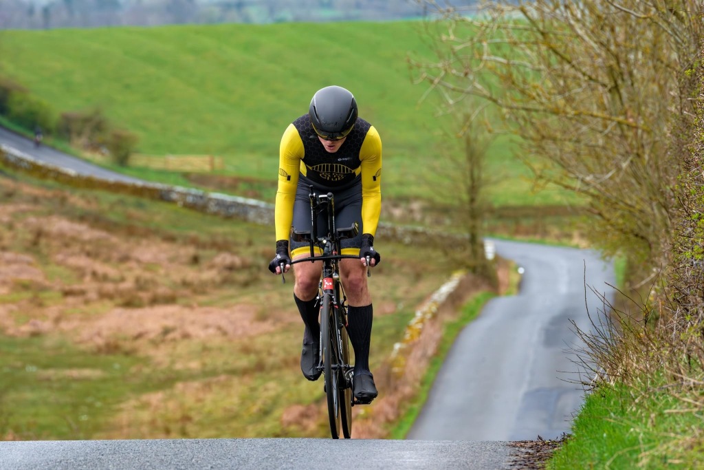 Cyclist riding a time trial bike in the base bar up the bank past Rothley Farm. Wearing the Tyne Triathlon Club skinsuit and a TT helmet.